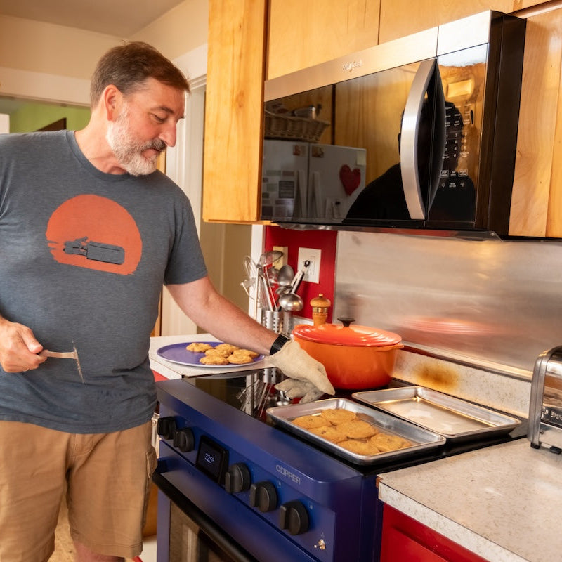 A man in a kitchen wearing a gray t-shirt with an orange graphic and khaki shorts, standing at a blue Charlie stove by Copper and placing a plate of cookies into an oven tray.