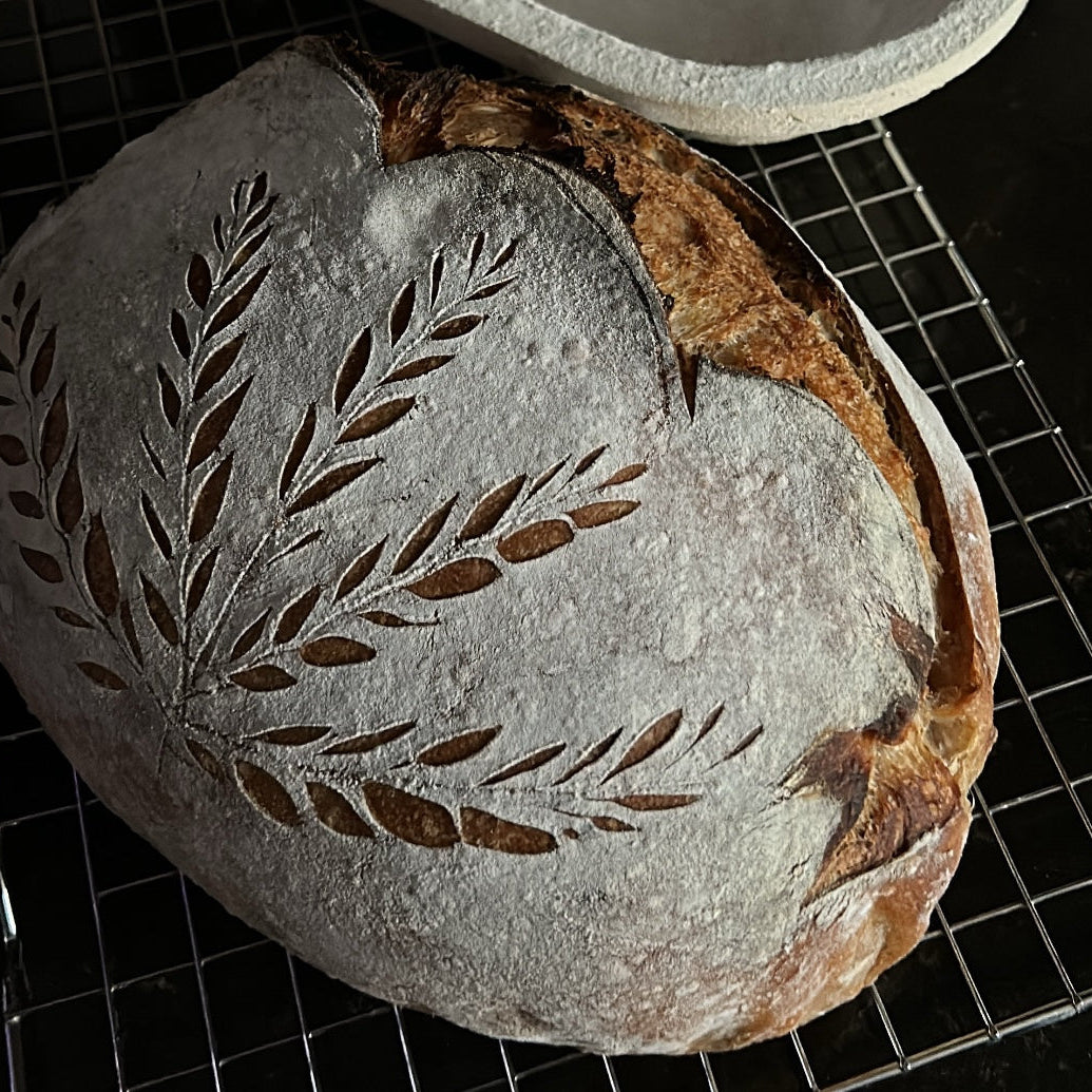 A round artisanal sourdough bread loaf with a flour-dusted crust and decorative leaf cutouts, resting on a black wire cooling rack.