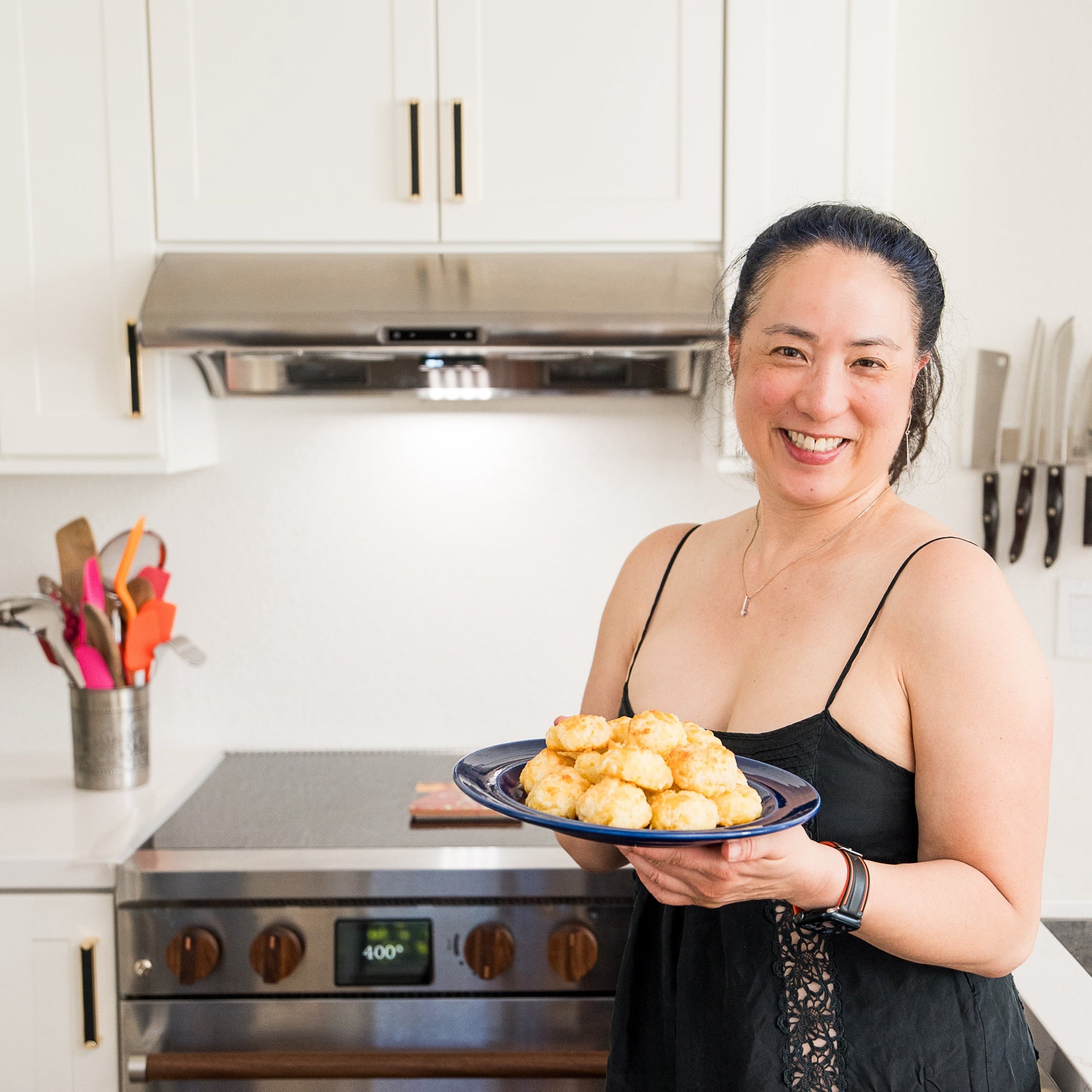 A smiling woman in a black spaghetti dress stands in a bright white kitchen, holding a blue plate filled with golden-brown buscuits. Behind her is a Copper Charlie battery-equipped induction range set to 400 degrees.