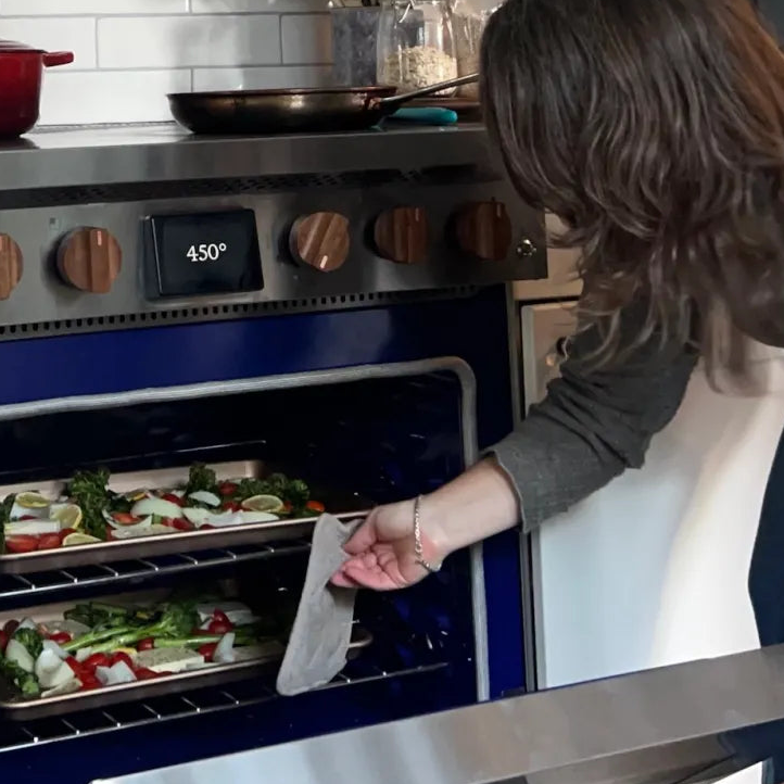 A woman checks the trays of vegetables she is roasting at 450° in her battery-equipped induction Copper Charlie range. The range features walnut wood knobs. The vegetables are broccolini, peppers, and onions with blocks of feta cheese and lemon spread out on 2 baking trays, one on the top rack and one on the bottom.